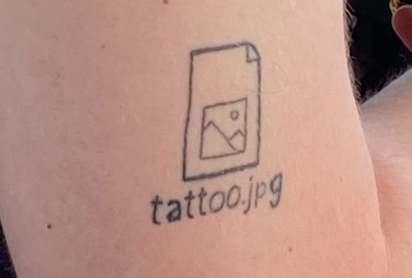 Eine Aufnahme eines Oberarms, darauf ein Tattoo von einem Computericon mit Beschriftung „tattoo.jpg“
