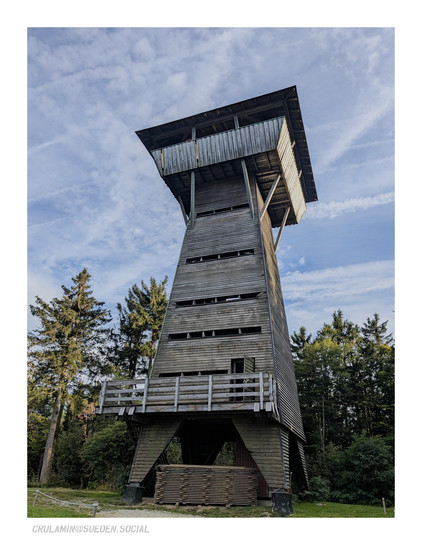 Ein leicht spitz zulaufender Aussichtsturm aus Holz, schon etwas verwittert und grau. Der Aufstieg erfolgt im Inneren, oben ist eine umlaufende, erweiterte Plattform mit Dach.Der Turm ist etwas über 20m hoch und schaut über die Spitzen der Fichten im Hintergrund. Darüber ist blauer Himmel mit Schleierwolken zu erkennen.