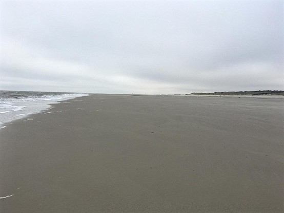 Ein Blick auf einen weiten, glatten, grauen Sandstrand bei Ebbe. Etwas oberhalb der Mitte liegt der Horizont. Der Himmel überzogen mit einer grauen verschwommenen Wolkendecke. Links ein Rest vom ablaufenden Wasser. Rechts am Horizont ein kleiner Streifen Dünen.

©️UR-F