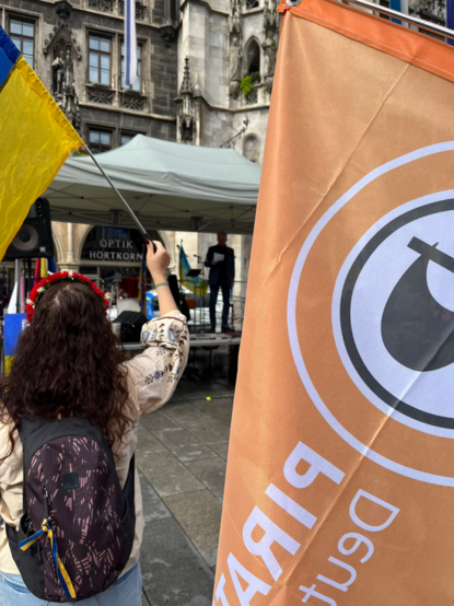 Ein Foto aus dem Publikum mit Blick auf die Tribüne, dort steht ein Redner.
Links im Bild eine Ukrainerin mit einer Ukraine-Flagge, rechts daneben, die Piratenpartei Flagge in Orange.